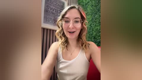 Media: Video of a smiling young woman with wavy blonde hair, wearing glasses and a white tank top, standing in a caf\u00e9 with greenery and a chalkboard menu in the background.