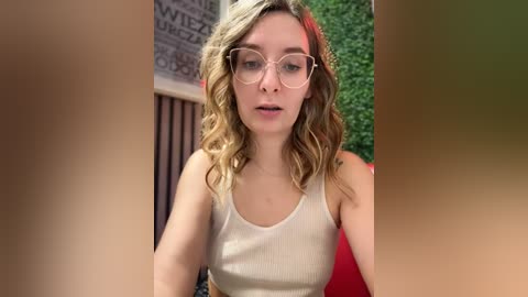 Media: Video of a young, light-skinned woman with wavy, blonde hair, wearing glasses, a beige tank top, and a neutral expression, seated in a cozy indoor setting with greenery and a chalkboard wall in the background.