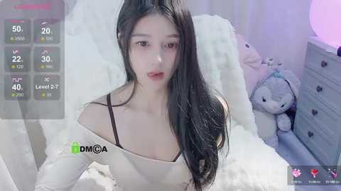 Media: Video of an East Asian woman with long black hair, light skin, wearing a white off-shoulder top, in a soft-focus, plush-filled room with a teddy bear.