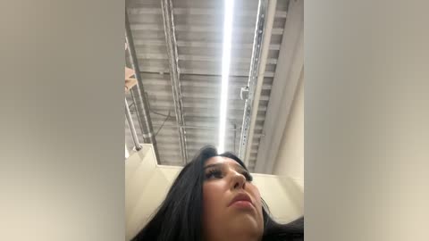 Media: Video of a woman with long, straight black hair, light skin, and full lips, lying on her back on a white bed. The ceiling is metallic with exposed pipes and a fluorescent light fixture. The image has a candid, unposed feel.