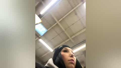 Media: Video of a young woman in a black beanie, staring up at fluorescent-lit ceiling in a dimly lit room. Her facial expression is neutral, and she wears a dark jacket.