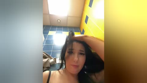 Media: A video of a woman with long black hair, light skin, and medium breasts, sitting in a bathroom with blue-tiled walls and yellow accents. She wears a black bra.