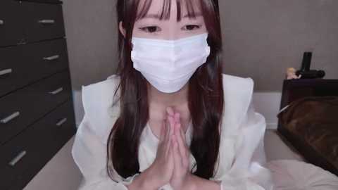 Media: Video of an Asian woman with long dark hair, wearing a white surgical mask, white lab coat, and praying with hands clasped in a hospital setting.