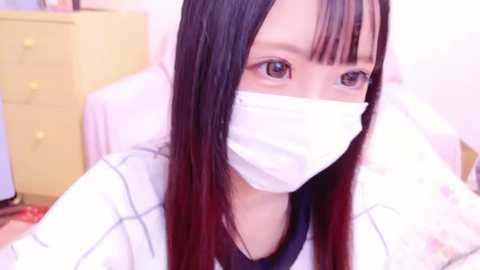 Media: Video of an East Asian woman with long, straight black hair, wearing a white mask, navy blue shirt, and a white robe. Background includes a yellow dresser and soft, pastel-colored walls.