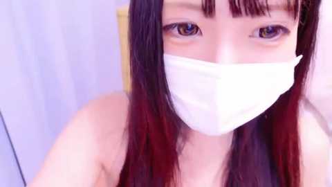 Media: Video of an Asian woman with long, straight black hair and a white surgical mask covering her mouth and nose. She has fair skin and light brown eyes.