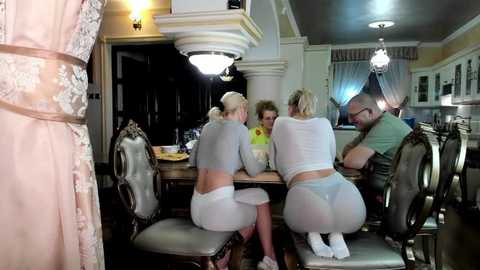 Media: Video of a stylish, well-lit dining room with four people, three women in white lingerie and one man in casual clothes, seated at a wooden table with ornate chairs.