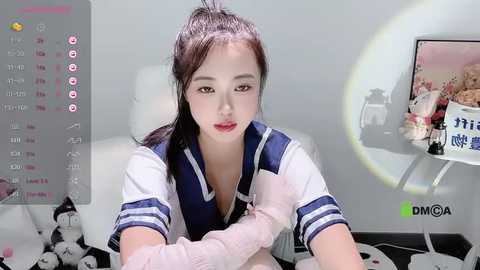 Media: Video of an Asian woman with fair skin, wearing a sailor schoolgirl uniform, seated on a white chair. Background features a white wall with a decorative lamp and plush toys.