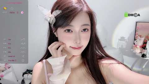 Media: A video of an East Asian woman with long, straight dark hair and a white headband, wearing a pink bra. She has fair skin, light makeup, and is smiling. The background features a minimalistic, white room with a computer screen displaying a calendar.