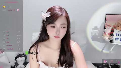 Media: Video of a young East Asian woman with long dark hair and a white flower in her hair, wearing a white lace bralette. Background features a bed with a teddy bear and a large circular mirror.