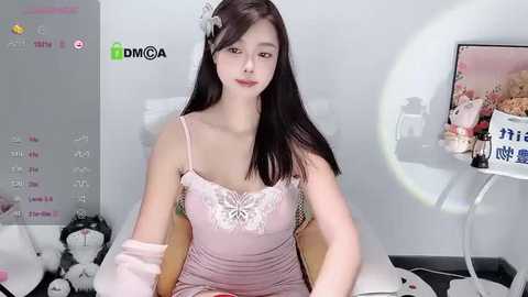 Media: Video of a young Asian woman with long black hair, wearing a pink lace camisole, sitting in a room with a white wall, a large round mirror, and a white bed.