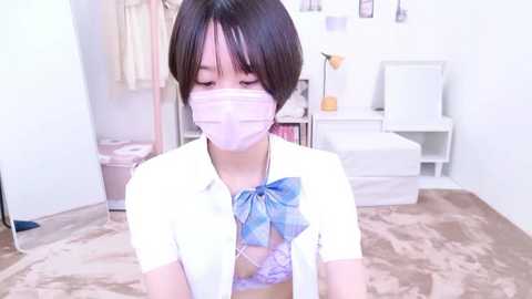 Media: Video of a petite, fair-skinned Asian woman with short black hair, wearing a white blouse, blue bow, and pink face mask, seated on a beige rug in a bright, minimalist bedroom.