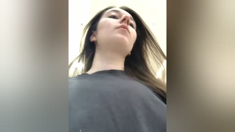 Media: A low-angle video of a young woman with long, straight brown hair, wearing a black t-shirt, standing in an indoor setting with soft, diffused lighting.