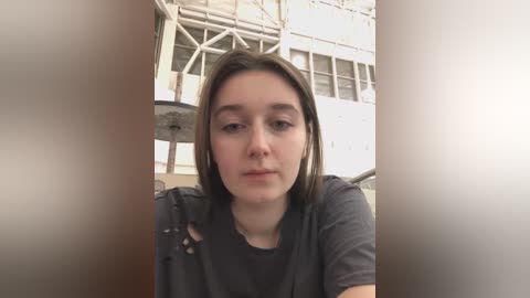 Media: A video of a young woman with straight, shoulder-length brown hair, wearing a dark t-shirt, indoors, with blurred background featuring industrial elements and a metal structure.