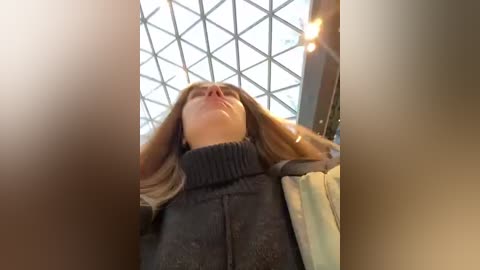 Media: Video of a woman with long brown hair, wearing a dark sweater, standing in a modern building with a glass ceiling and bright lighting, giving a tilted, low-angle view.