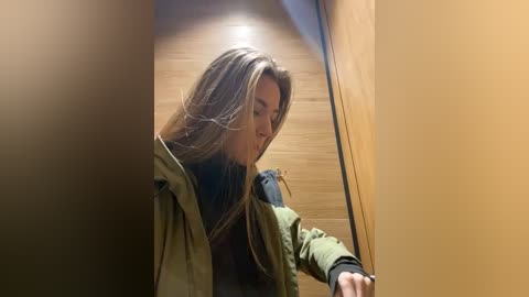 Media: A video of a woman with long, light brown hair in a beige jacket, leaning against a wooden door in a dimly lit hallway.