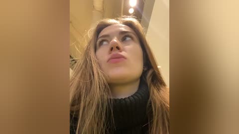 Media: A video of a young woman with light skin and long, wet, brown hair, wearing a black turtleneck sweater, gazing upward with a pensive expression. The background is blurred, featuring warm lighting and an indistinct indoor setting.