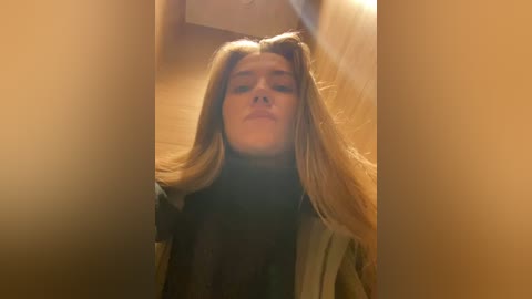 Media: A video of a young woman with long blonde hair, wearing a black turtleneck and beige coat, standing in a dimly lit hallway with warm, golden light.