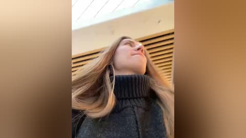 Media: Video of a Caucasian woman with long, light brown hair, wearing a dark gray turtleneck sweater, standing in a narrow, beige-walled hallway with wooden slatted blinds.