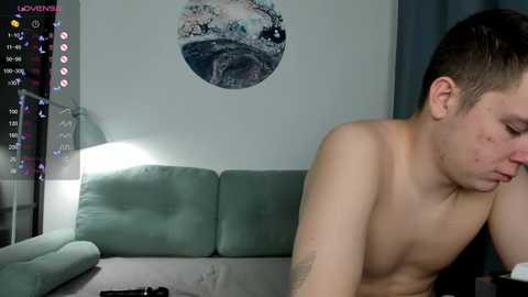 Media: Video of a shirtless, slim, young man with short dark hair, sitting on a green couch in a dimly lit room with a moon phase painting on the wall.