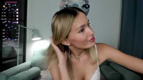 Media: Video of a smiling, blonde-haired woman with light skin, wearing a white lace camisole, adjusting her hair. She's in a modern living room with a blue sofa, a white lamp, and a moon-shaped wall decoration.