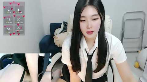 Media: Video of a young Asian woman with long black hair, wearing a white shirt and black tie, sitting on a bed with a plush toy, in a simple room with a calendar.