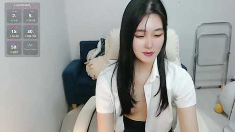 Media: Video of an Asian woman with long black hair, wearing a white blouse, sitting on a beige chair in a minimalistic room with a stuffed bear and a digital display.