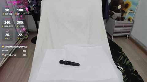 Media: Video of a reclining chair draped with white fabric, featuring a black dildo on the cushion. The background includes a teddy bear and floral artwork. Live video overlay shows viewers' comments and stats.