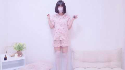 Media: Video of a young Asian woman with a short bob haircut and fair skin, wearing a pink pajama set and face mask, standing in a minimalist room with white walls, a white couch, and a potted plant.