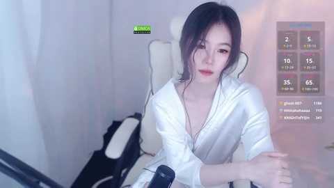 Media: A video of an Asian woman with fair skin, wearing a white bathrobe, sitting in a white gaming chair, with a gaming overlay showing stats and settings.