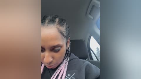 Media: Video of a young Black woman with braided hair, nose ring, and pink headband, sitting in a car with a window view of trees.