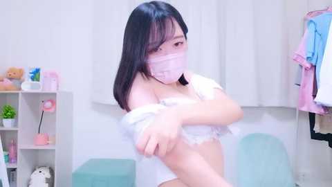 Media: Video of a young East Asian woman with long black hair, wearing a white lace bra, white face mask, and a white shirt, adjusting it in a brightly lit room with pastel decor, including shelves with toys and clothes.