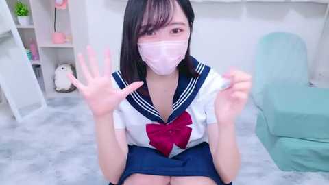 Media: Video of an Asian woman with pale skin, straight black hair, and a pink face mask, wearing a sailor schoolgirl outfit with a large red bow. She sits on a light-colored floor, smiling and holding up her hands.