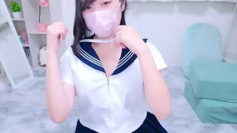 Media: Video of an Asian woman in a pink mask, white sailor collar blouse, and navy skirt, posing playfully in a pastel-themed room with a green chair and white shelves.