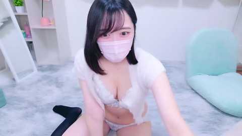 Media: Video of an East Asian woman with shoulder-length black hair, wearing a white lace bra and panties, a pink mask, and a white jacket, kneeling on a gray carpet in a minimalist room with white walls and light-colored furniture.