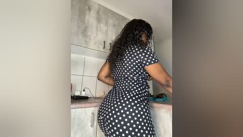 Media: Video of a dark-skinned woman with curly hair in a black polka-dot dress, standing in a minimalist kitchen with white tiles, a sink, and a countertop.