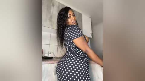 Media: Video of a confident Black woman with dark skin and long curly black hair, wearing a black polka dot dress, standing in a modern kitchen with white cabinets and gray countertops.