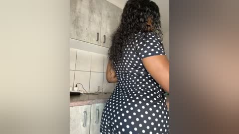 Media: Video of a dark-skinned woman with long curly hair, wearing a black polka-dot dress, standing in a minimalist kitchen with white tiles and gray cabinets.