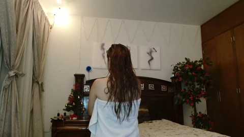 Media: Video of a woman with long, wet, brown hair, wearing a white towel, standing in a dimly lit bedroom with a wooden bed, nightstands, and floral decorations.