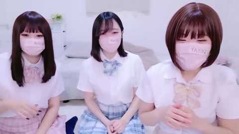 Media: Video of three East Asian women in white school uniforms, wearing face masks, sitting in a bright, minimalist room with a grey couch and flowers.