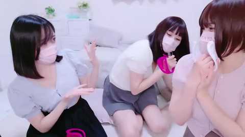 Media: Video of three Asian women wearing surgical masks, white gowns, and gloves, kneeling on a white bed, discussing something, in a sterile, brightly lit room.