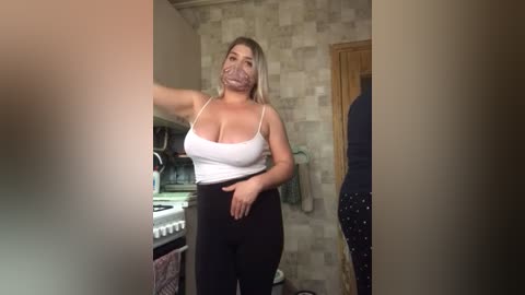 Media: Video of a curvy, blonde woman in a white tank top and black pants, standing in a beige-tiled kitchen, with a partially visible person in the background.