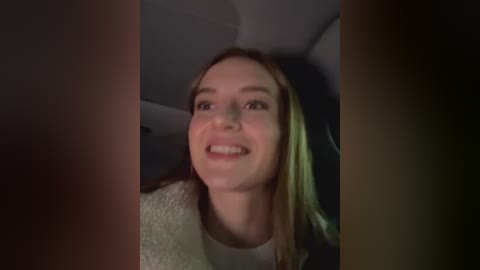 Media: A video of a young woman with fair skin and straight brown hair, smiling, seen from a slightly low angle, sitting in a dimly lit car interior.