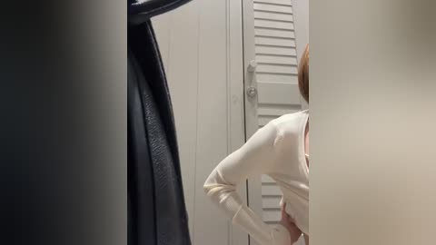 Media: Video of a person in a beige sweater, facing right, partially obscured by a black coat, standing in front of white shuttered doors.