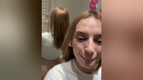 Media: A video of a young woman with light skin and shoulder-length blonde hair, wearing a white shirt, standing in a bathroom. She smiles, looking at her reflection in the mirror. The background includes a pink \"Hair\" sign.