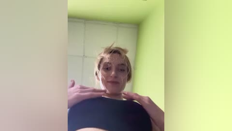 Media: A video of a fair-skinned woman with blonde hair, wearing a black sports bra, standing in a brightly lit room with green walls and white tiled ceiling.