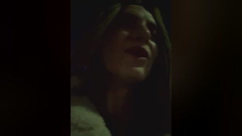 Media: Video of a woman's partially visible face with dark hair, wearing a hooded cloak, in a dimly lit, dark room, creating a mysterious, ominous atmosphere.