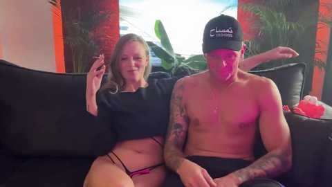 Media: Video of a topless, tattooed man in a black cap and a woman in a black crop top and pink panties, sitting on a black couch with a tropical background.