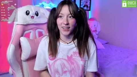 Media: A young woman with straight brown hair in pigtails, wearing a white T-shirt with a pink graphic, sits in a plush, pink gaming chair in a softly lit room with anime-themed decor.