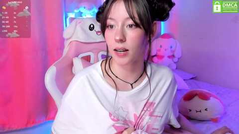 Media: Video of a young woman with dark hair in pigtails, wearing a white shirt, sitting in a pink chair with plush toys, in a room with pastel colors and a playful atmosphere.