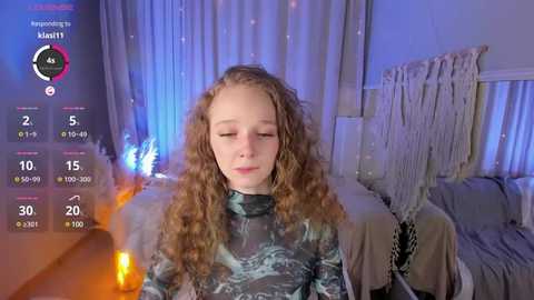 Media: Video of a young Caucasian girl with curly brown hair, wearing a camo-patterned long-sleeve shirt, sitting in a dimly lit bedroom with a bed and white curtains in the background.